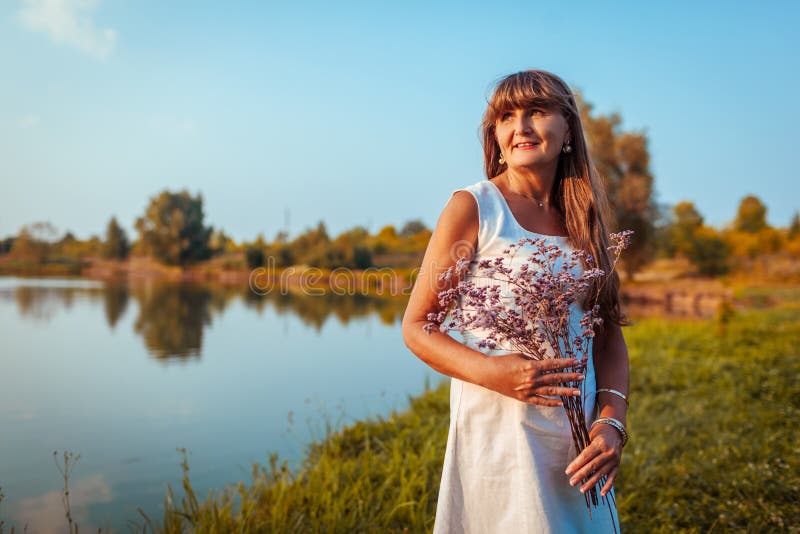 Mature Woman Holding Flowers In Front Of The River In Autumn Park Senior Woman Admires