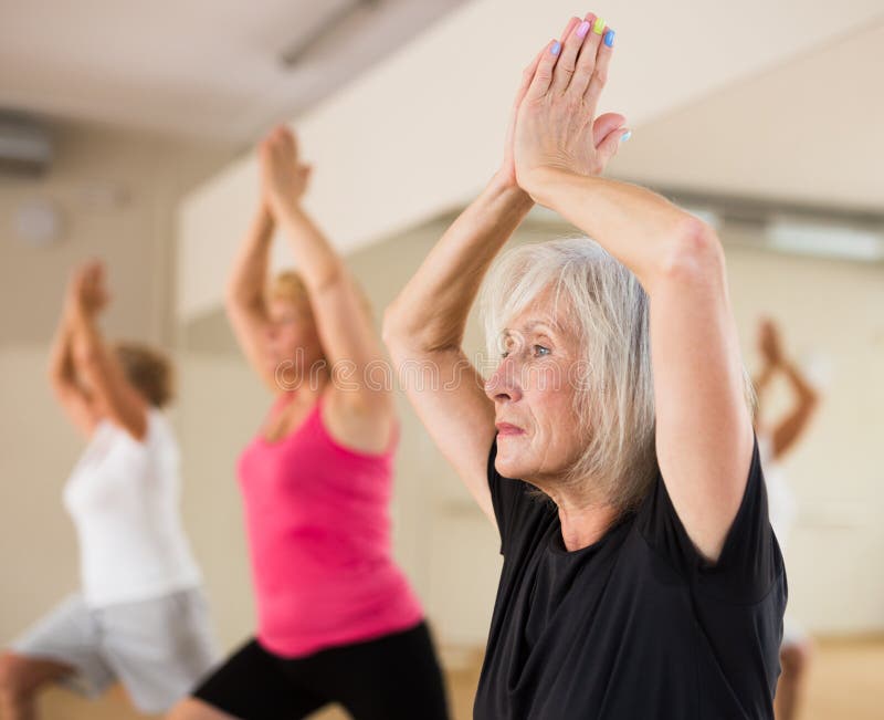 Mature women practicing yoga at a group training session in a fitness studio perform an exercise in the warrior pose I. Mature women practicing yoga at a group training session in a fitness studio perform an exercise in the warrior pose I