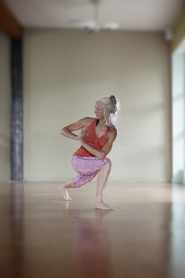 Mature yogini practicing the Warrior Twist Pose. She is wearing a pretty orange and pink yoga outfit while practicing in a beautiful studio with a wood floor. Mature yogini practicing the Warrior Twist Pose. She is wearing a pretty orange and pink yoga outfit while practicing in a beautiful studio with a wood floor.