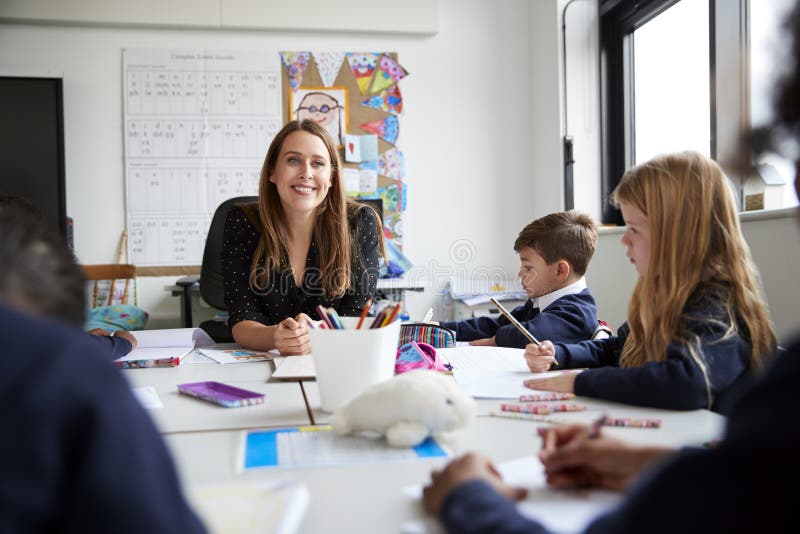 Maître d'école primaire féminin s'asseyant à une table souriant à la caméra pendant une leçon avec un groupe d'écoliers, angle fa photos libres de droits Maître d'école primaire féminin s'asseyant à une table souriant à la caméra pendant une leçon avec un groupe d'écoliers, angle fa photos libres de droits