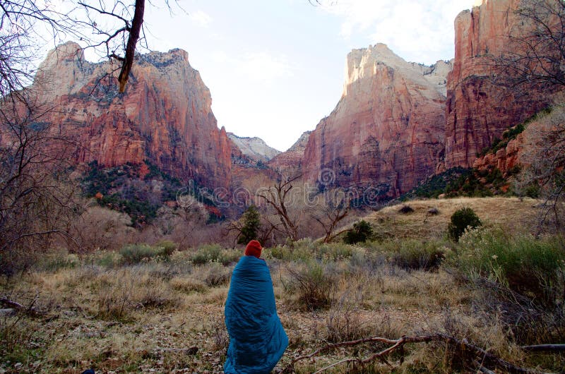 Early in the morning, a young woman wrapped in a sleeping bag wakes up to the beautiful panorama of the mountains in Zion National Park. Early in the morning, a young woman wrapped in a sleeping bag wakes up to the beautiful panorama of the mountains in Zion National Park