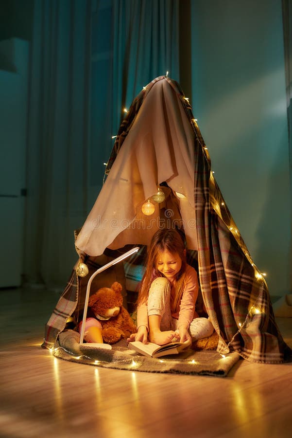 A little girl wearing pyjamas sitting on a floor barefoot in a self-made hut made of a plaid, enjoying her time reading a book with fancy garlands all around the hut in a cosy big darken room. A little girl wearing pyjamas sitting on a floor barefoot in a self-made hut made of a plaid, enjoying her time reading a book with fancy garlands all around the hut in a cosy big darken room