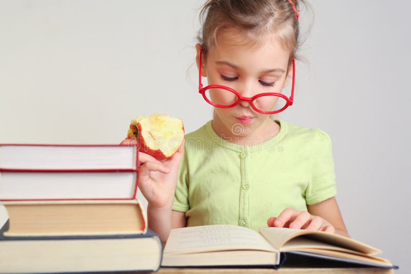 Little girl in glasses eat apple, read book near stack of books. Little girl in glasses eat apple, read book near stack of books