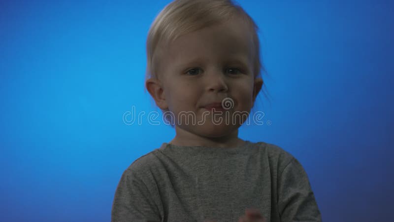 Małe blond kaukaskie dziecko w szarym koszulku uśmiecha się i pokazuje kciuki w kamerze i odchodzi