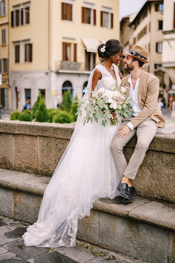 African-American bride in a white dress with a long veil and bouquet, and Caucasian groom in a sand jacket and straw hat. Interracial wedding couple. Wedding in Florence, Italy. African-American bride in a white dress with a long veil and bouquet, and Caucasian groom in a sand jacket and straw hat. Interracial wedding couple. Wedding in Florence, Italy.