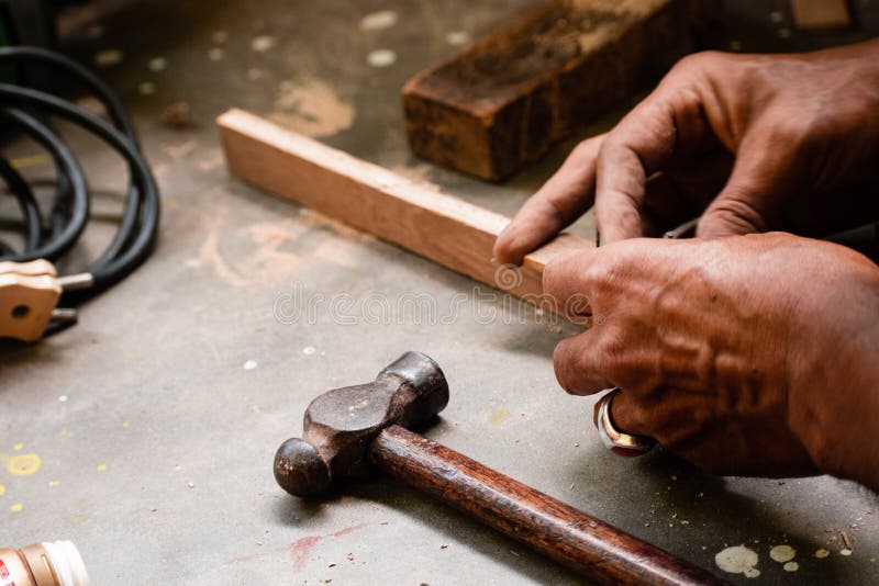 A mechanic does carpenter jobs with wood, plus and other tools. He is hitting hammer on the head of nail to wooden pieces and cutting wooden sticks by saw. A drill machine is kept on floor beside him