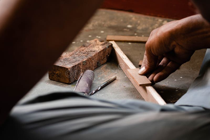 A mechanic does carpenter jobs with wood, plus and other tools. He is hitting hammer on the head of nail to wooden pieces and cutting wooden sticks by saw. A drill machine is kept on floor beside him