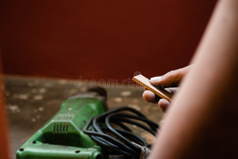 A mechanic does carpenter jobs with wood, plus and other tools. He is hitting hammer on the head of nail to wooden pieces and cutting wooden sticks by saw. A drill machine is kept on floor beside him