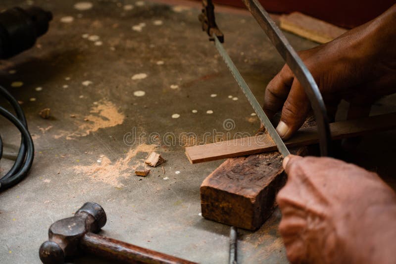 A mechanic does carpenter jobs with wood, plus and other tools. He is hitting hammer on the head of nail to wooden pieces and cutting wooden sticks by saw. A drill machine is kept on floor beside him