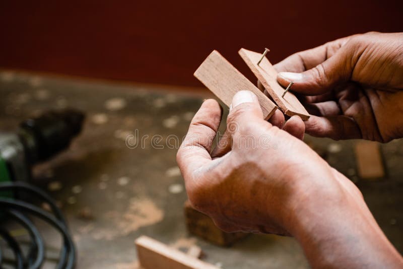 A mechanic does carpenter jobs with wood, plus and other tools. He is hitting hammer on the head of nail to wooden pieces and cutting wooden sticks by saw. A drill machine is kept on floor beside him