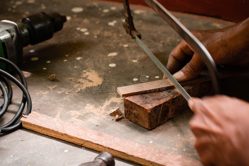 A mechanic does carpenter jobs with wood, plus and other tools. He is hitting hammer on the head of nail to wooden pieces and cutting wooden sticks by saw. A drill machine is kept on floor beside him