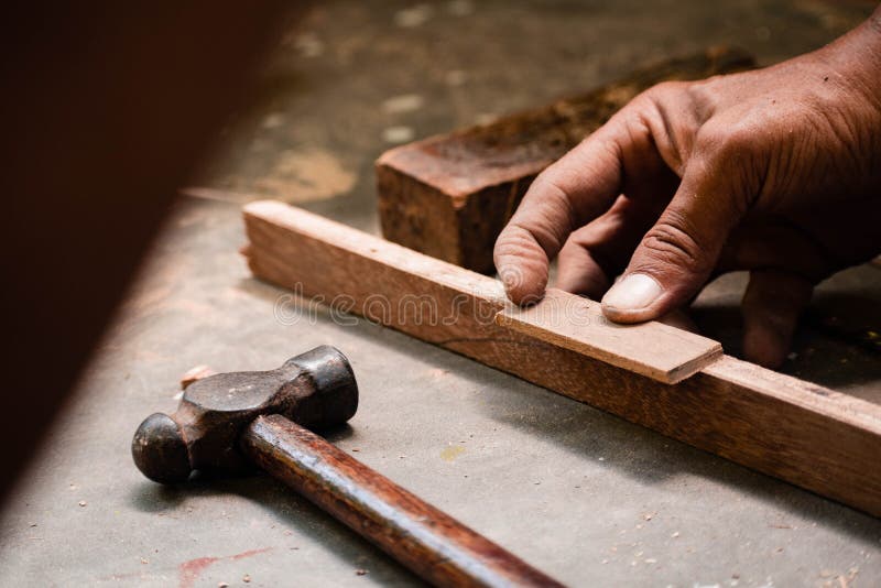 A mechanic does carpenter jobs with wood, plus and other tools. He is hitting hammer on the head of nail to wooden pieces and cutting wooden sticks by saw. A drill machine is kept on floor beside him
