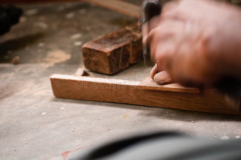 A mechanic does carpenter jobs with wood, plus and other tools. He is hitting hammer on the head of nail to wooden pieces and cutting wooden sticks by saw. A drill machine is kept on floor beside him