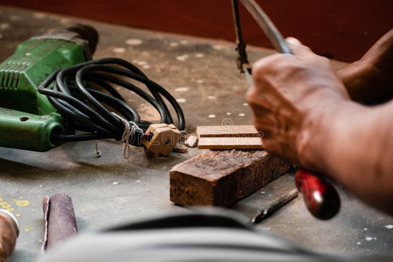 A mechanic does carpenter jobs with wood, plus and other tools. He is hitting hammer on the head of nail to wooden pieces and cutting wooden sticks by saw. A drill machine is kept on floor beside him