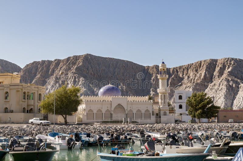 Meczet Sceniczny w Omanu Khasabie i Widok na Port. Khasab Oman przedstawia malowniczą scenę z meczetem charakteryzującym się uderzającą niebieską kopułą i wysokim minaretem. Zagnieżdżony na chropowatym terenie górskim, na pierwszym planie znajduje się port wypełniony licznymi łodziami o wysokich korzeniach pływającymi po spokojnej wodzie. Sąsiadujące budynki wykazują tradycyjne elementy architektoniczne harmonizujące się z meczetem. Drzewa łuskane kropkowane krajobraz dodając dotknięcia zieleni. Tło skalistych gór wzmacnia piękno tego wybrzeża.