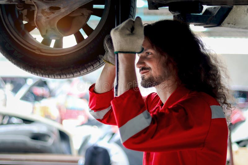 Mecánico de pelo largo y apuesto hombre con uniforme rojo trabajo con vehículo levantado motomecánico rueda de control en el técni imagen de archivo
