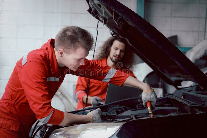 Mecánico de uniforme rojo cargador batería de coche con electricidad a través de cables puente. compañeros de trabajo trabajan jun imagen de archivo libre de regalías