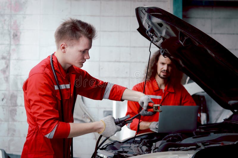 Mecánico de uniforme rojo cargador batería de coche con electricidad a través de cables puente. compañeros de trabajo trabajan jun foto de archivo libre de regalías