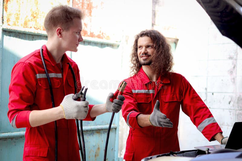 Mecánico de uniforme rojo cargador batería de coche con electricidad a través de cables puente. compañeros de trabajo trabajan jun imagenes de archivo
