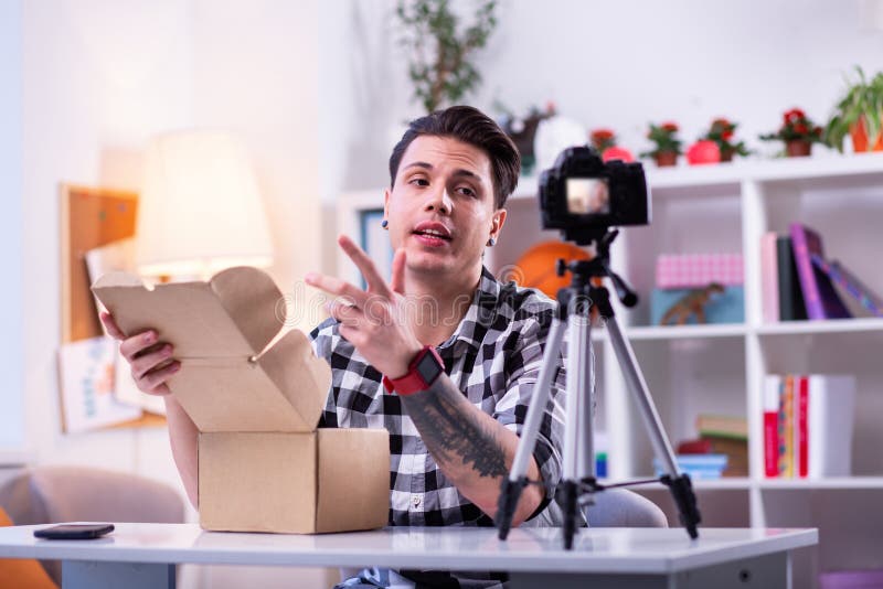 Beaming young man. Communicative dark-haired guy in checkered shirt unboxing package and recording process on camera. Beaming young man. Communicative dark-haired guy in checkered shirt unboxing package and recording process on camera