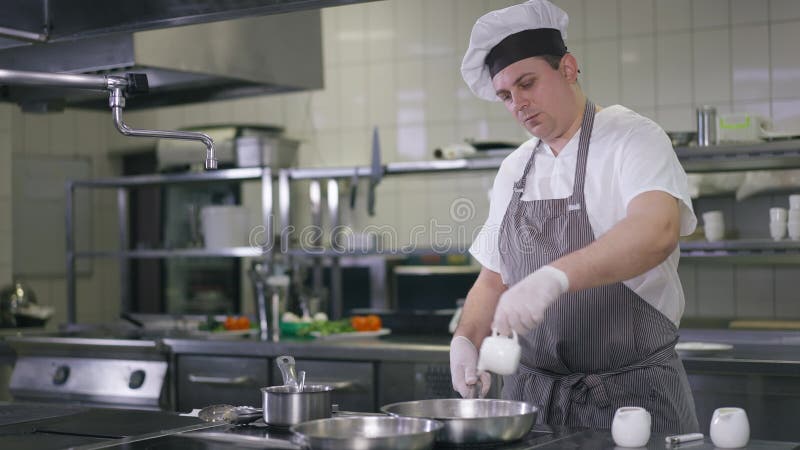 Medel-shot självsäker proffskkock i kock som häller vatten i pall-frys-middag i restaurangköket. allvarlig