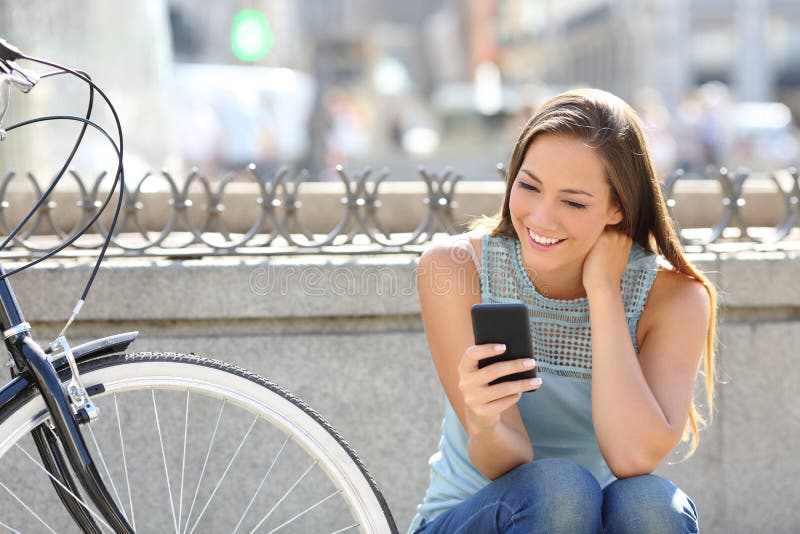 Happy girl watching media in a smart phone sitting in the street beside her bicycle. Happy girl watching media in a smart phone sitting in the street beside her bicycle