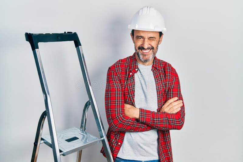Mediana edad hispano trabajador hombre con los brazos cruzados gesto usando escalera guiñando la mirada a la cámara con expresión fotos de archivo