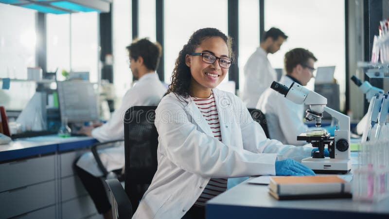 Medical Science Laboratory: Beautiful Black Scientist is Using Microscope, Looking at Camera and stock image
