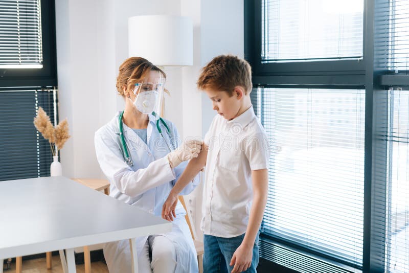 Medico donna con uniforme bianca e maschera di sicurezza che applica gesso sulla spalla di un bambino dopo la vaccinazione immagine stock libera da diritti