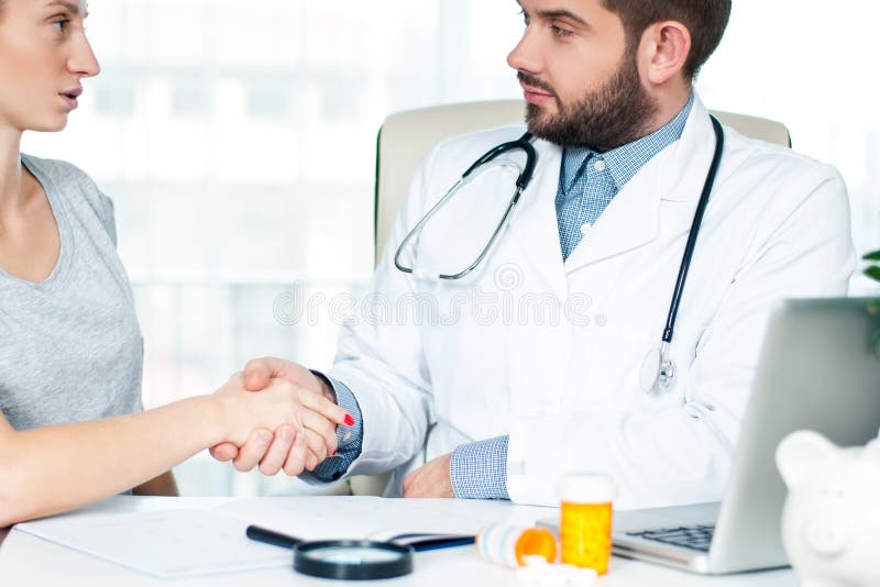 Medico sorridente che stringe le mani con un paziente femminile nell'ufficio fotografia stock Medico sorridente che stringe le mani con un paziente femminile nell'ufficio fotografia stock