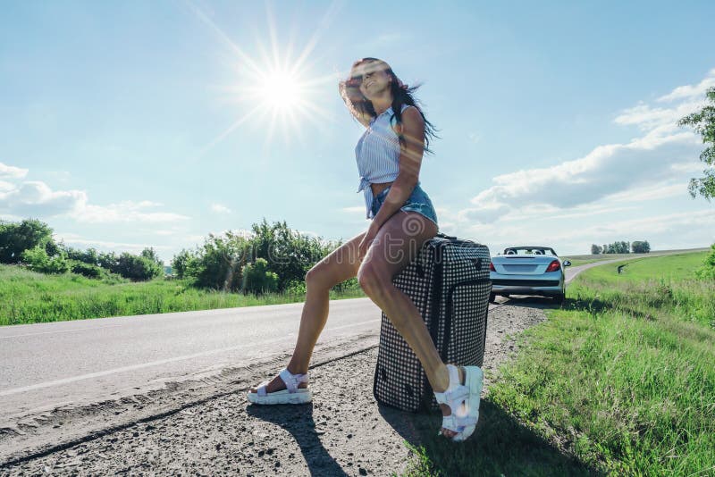 Meisje in de heldere zon op weg met Bagage op de achtergrond van de auto Kaukasische vrouwenzitting op een koffer E stock afbeeldingen Meisje in de heldere zon op weg met Bagage op de achtergrond van de auto Kaukasische vrouwenzitting op een koffer E stock afbeeldingen