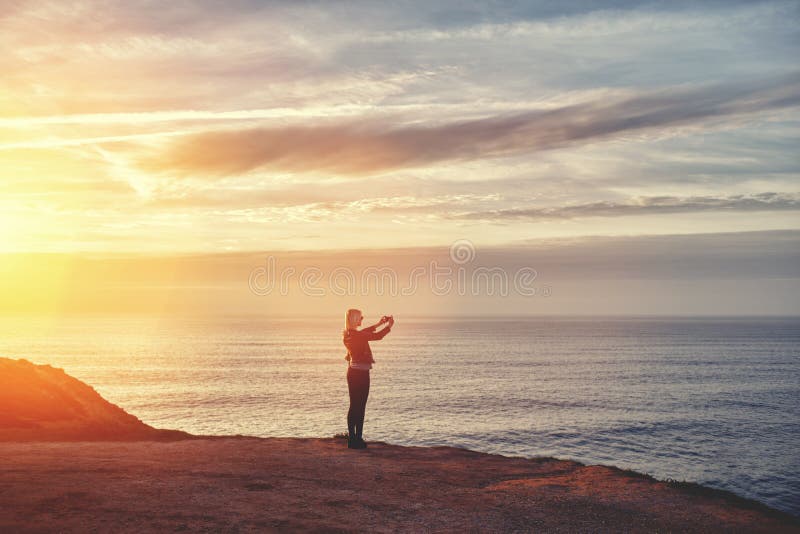 Meisje die foto met mobiele telefooncamera nemen terwijl status op bergheuvel tegen verbazende oceaan en zonsondergangachtergrond stock afbeeldingen Meisje die foto met mobiele telefooncamera nemen terwijl status op bergheuvel tegen verbazende oceaan en zonsondergangachtergrond stock afbeeldingen