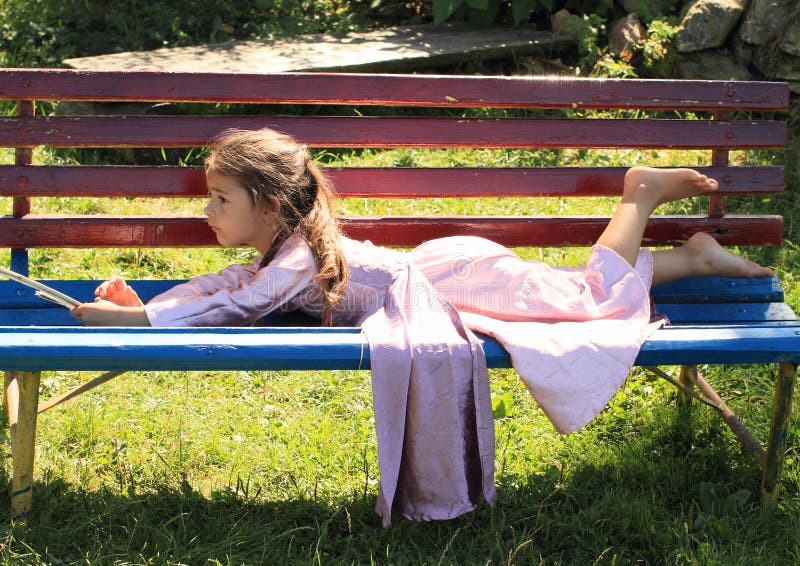 Barefoot girl in lila dress lying on red and blue bench and having rest. Barefoot girl in lila dress lying on red and blue bench and having rest