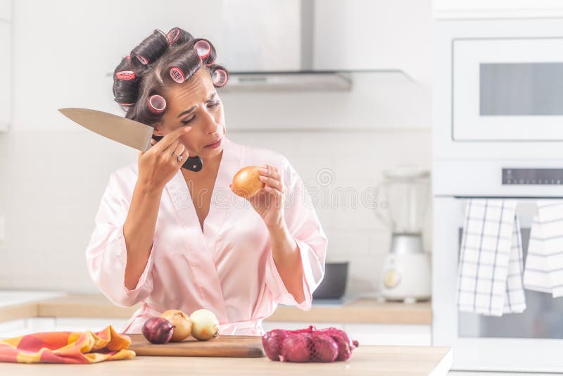 Girl cries over onion chopping in the kitchen wearing head curlers in the kitchen. Girl cries over onion chopping in the kitchen wearing head curlers in the kitchen