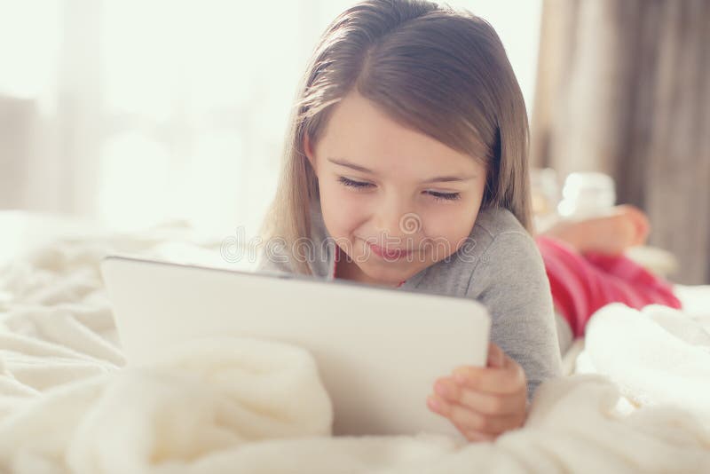 The nice little girl of 6 years, the brunette with soft hair, in pink striped pajama trousers and a gray t-shirt, lying on the bed covered with a fluffy white cover communicates with friends on social networks, using the tablet computer of white color. The nice little girl of 6 years, the brunette with soft hair, in pink striped pajama trousers and a gray t-shirt, lying on the bed covered with a fluffy white cover communicates with friends on social networks, using the tablet computer of white color