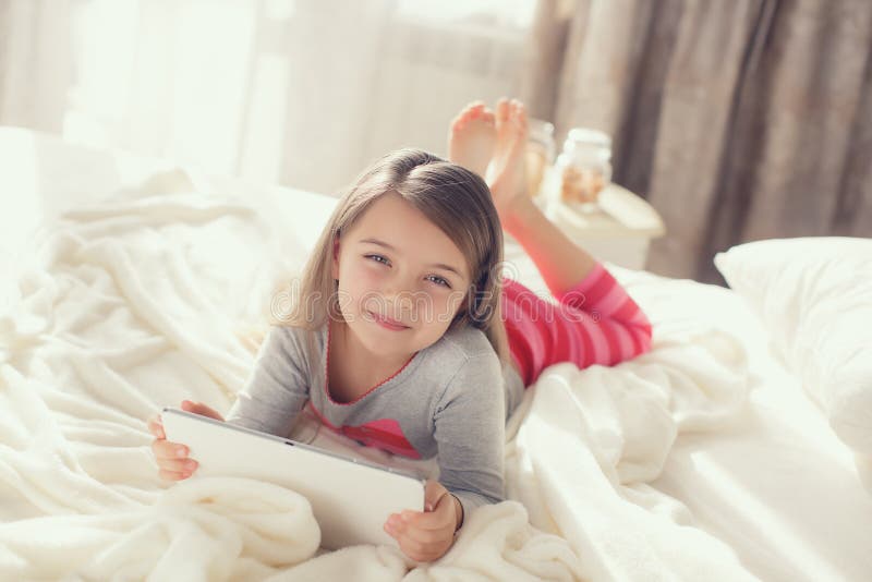 The nice little girl of 6 years, the brunette with soft hair, in pink striped pajama trousers and a gray t-shirt, lying on the bed covered with a fluffy white cover communicates with friends on social networks, using the tablet computer of white color. The nice little girl of 6 years, the brunette with soft hair, in pink striped pajama trousers and a gray t-shirt, lying on the bed covered with a fluffy white cover communicates with friends on social networks, using the tablet computer of white color