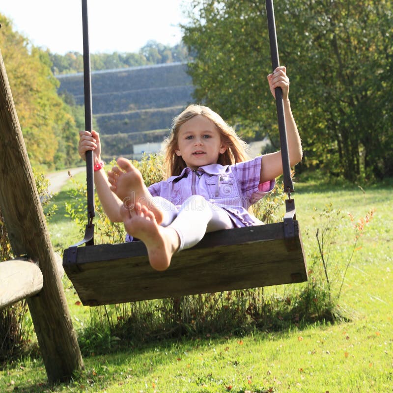 Barefoot kid - smiling girl in lila clothes sitting and swinging on a swing. Barefoot kid - smiling girl in lila clothes sitting and swinging on a swing