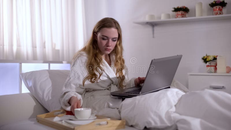 Meisje in robe met koffiekop in bed die aan laptop, freelance concept, baan werken stock afbeelding Meisje in robe met koffiekop in bed die aan laptop, freelance concept, baan werken stock afbeelding