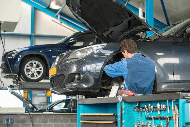 Mechanic, at work on the wheel hub and brake disk of a car in a garage, with other vehicles on car lifts in the background and a trolly with various tools, clamps and wrenches in the foreground. The hood of the car is open. Mechanic, at work on the wheel hub and brake disk of a car in a garage, with other vehicles on car lifts in the background and a trolly with various tools, clamps and wrenches in the foreground. The hood of the car is open