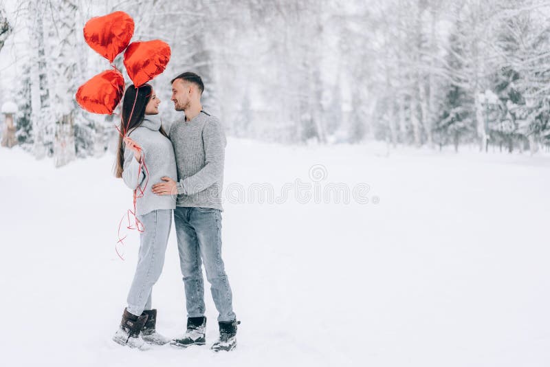 A man and a woman are hugging in a snowy park. A woman holds in her hands heart-shaped balloons filled with helium