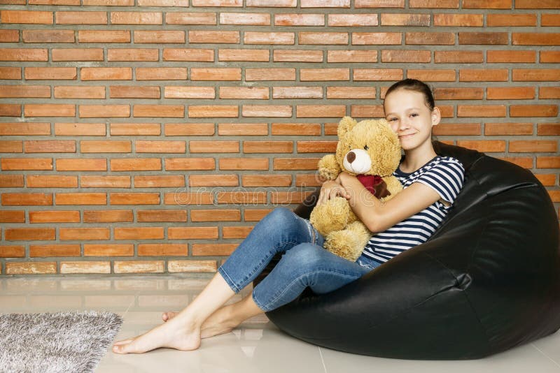 Menina adolescente caucasiano bonita que senta-se na cadeira do saco de feijão preto que guarda o brinquedo marrom grande do urso imagens de stock Menina adolescente caucasiano bonita que senta-se na cadeira do saco de feijão preto que guarda o brinquedo marrom grande do urso imagens de stock
