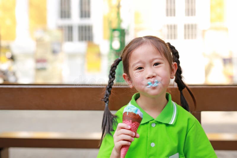 A menina asi?tica pequena de sorriso da crian?a aprecia comer o cone de gelado com manchas em torno de sua boca imagem de stock A menina asi?tica pequena de sorriso da crian?a aprecia comer o cone de gelado com manchas em torno de sua boca imagem de stock