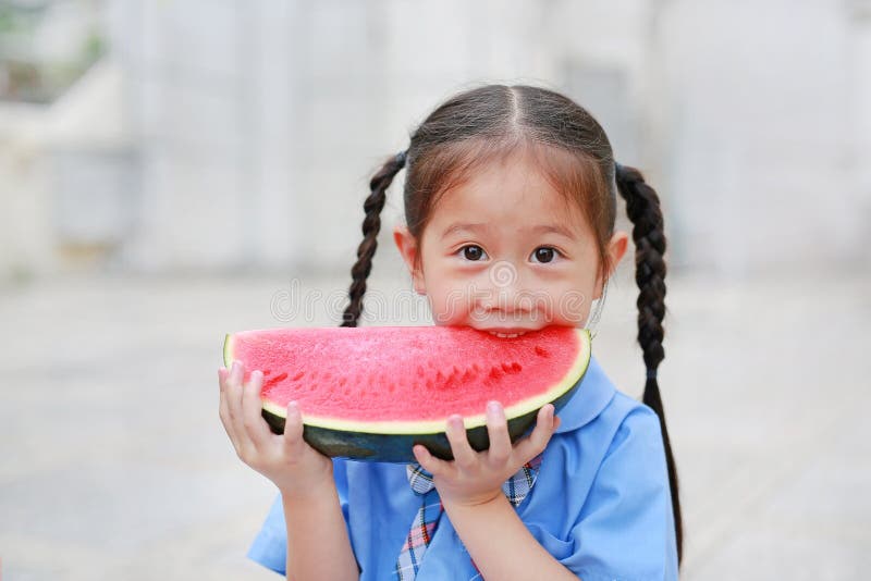 A menina asiática pequena bonito da criança na farda da escola aprecia comer a melancia cortada fresca fotografia de stock royalty free A menina asiática pequena bonito da criança na farda da escola aprecia comer a melancia cortada fresca fotografia de stock royalty free