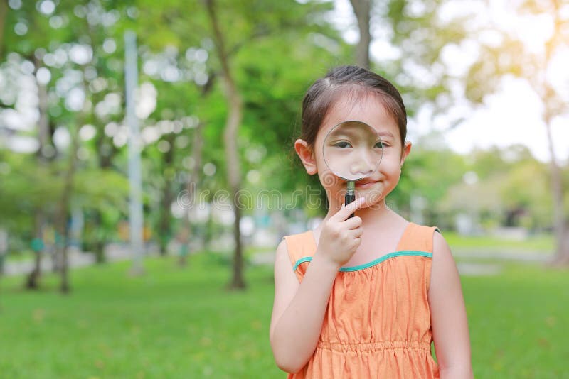 Menina asiática pequena da criança do retrato que olha através da lupa no jardim do parque imagens de stock Menina asiática pequena da criança do retrato que olha através da lupa no jardim do parque imagens de stock