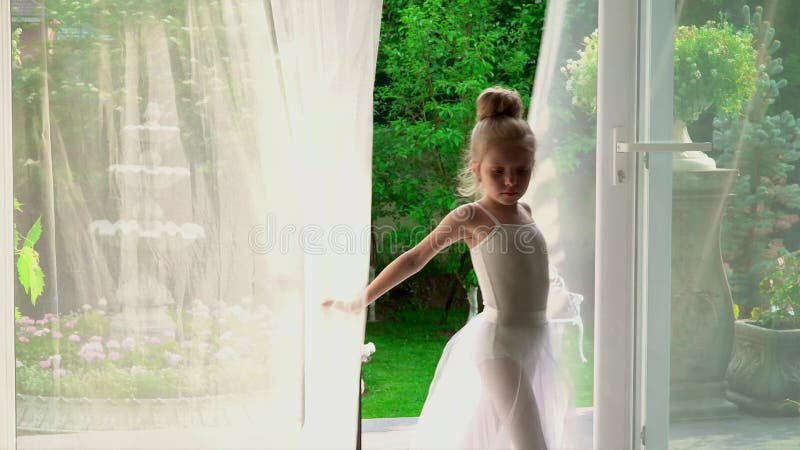 A menina a bailarina é pela janela A menina bonita em um tutu branco é pela janela de uma escola do bailado