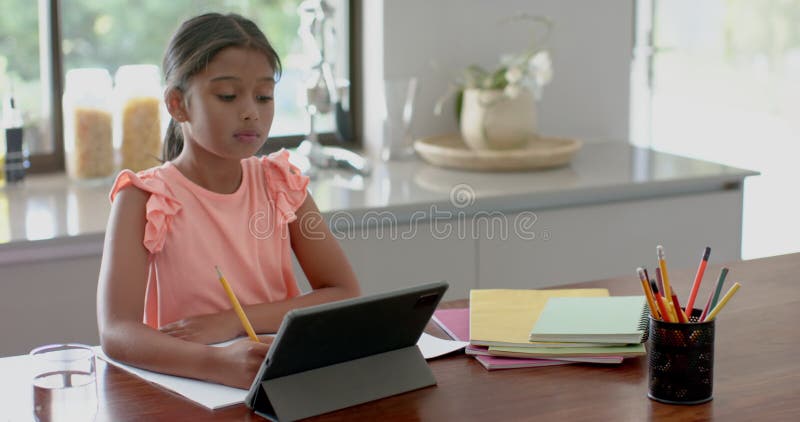 Menina biracial focada tendo aula primária em tablet na cozinha espaço em câmera lenta