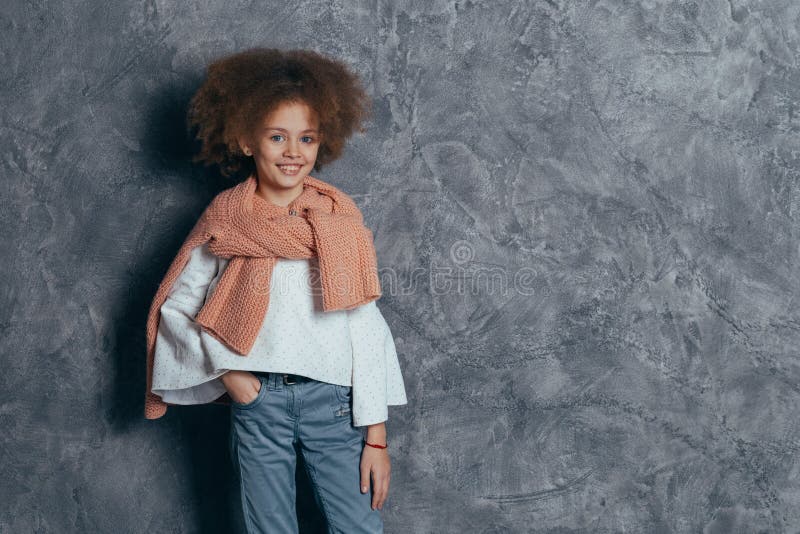 Menina bonita de sorriso com o cabelo encaracolado que está no estúdio na parede cinzenta imagem de stock Menina bonita de sorriso com o cabelo encaracolado que está no estúdio na parede cinzenta imagem de stock