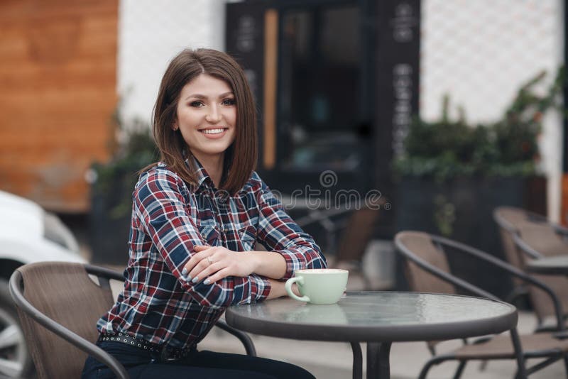 A menina bonita, morena bonita com penteado elegante em uma camisa de manta bebe a xícara de café fora no café do verão fotos de stock A menina bonita, morena bonita com penteado elegante em uma camisa de manta bebe a xícara de café fora no café do verão fotos de stock