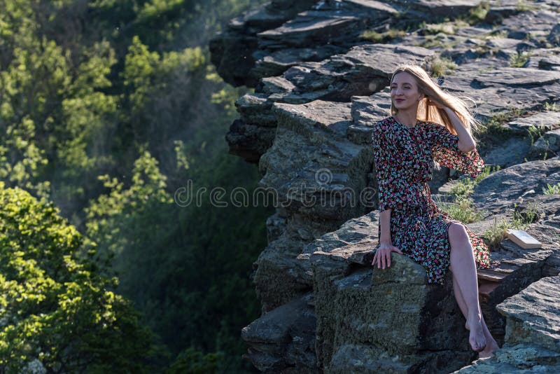 A menina bonita no vestido senta-se na rocha na natureza foto de stock A menina bonita no vestido senta-se na rocha na natureza foto de stock