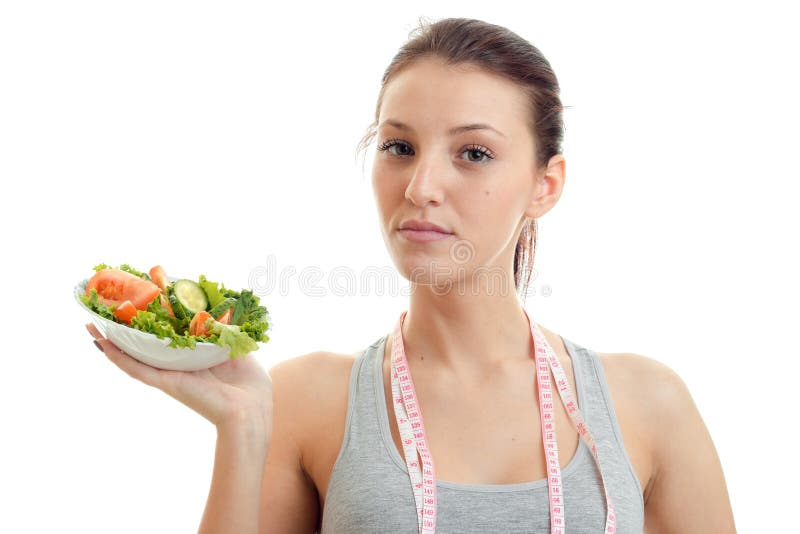 Young beautiful girl with the measuring tape around the neck looks into the camera and holds high in vegetable salad plate close-up isolated on white background. Young beautiful girl with the measuring tape around the neck looks into the camera and holds high in vegetable salad plate close-up isolated on white background.